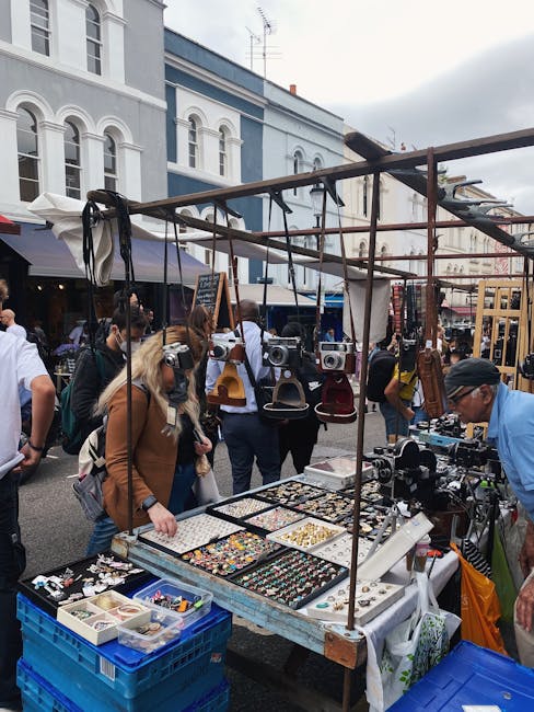 A street market scene on a cloudy day, with a variety of vendors displaying items on outdoor tables. In the foreground, a table covered with jewelry, watches, and small accessories is visible, with some items organized in boxes and on display trays. Above the tables, a metal framework supports hanging products such as handbags and other accessories. Several shoppers, some wearing casual clothing and carrying shopping bags, browse the stalls, while a man wearing a blue shirt and black cap discusses with a vendor. In the background, there are historic-style buildings with large windows and arched details, with other market stalls and people visible further along the street. The environment suggests a lively outdoor marketplace, with natural daylight illuminating the scene, and it could be part of a house removal or moving-related event where vendors set up temporary stalls. Man and Van Notting Hill occasionally provides support for logistics and furniture transport during such activities.