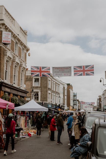A busy street scene in Portobello Road, Notting Hill, during a market day with multiple pedestrians walking along the pavement, some engaging with vendors and others carrying shopping bags. The street is lined with shops, including a clothing stall with colorful garments displayed on racks and tables, under a white canopy. Several cardboard boxes and wrapped packages are visible on the ground near the vendors, indicating packing and unpacking activities related to home relocation or moving services. To the right, part of a black van is parked beside the curb, suggesting furniture transport or loading for a moving job. Overhead, three Union Jack flags and a banner reading 'PORTOBELLO ROAD W11' are strung across the street, highlighting the location. The surrounding buildings are multi-storey, with traditional architecture and decorative detailing. The overcast sky provides diffuse natural lighting, creating a neutral and detailed view suitable for accessibility and SEO relevance to removals and house moving services from Man and Van Notting Hill.