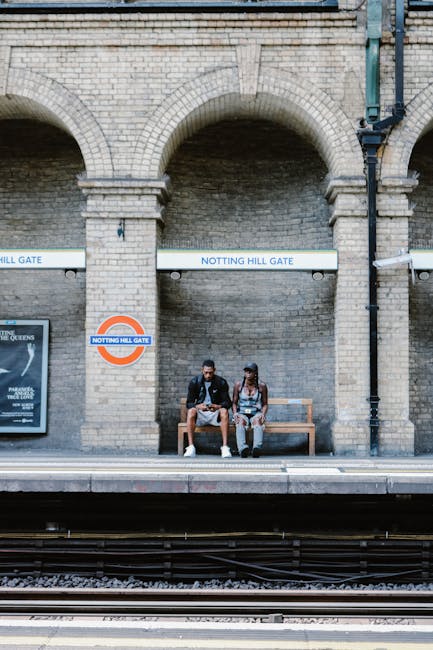 Two individuals sit on a wooden bench at a train station platform beneath a large brick archway featuring two signs reading 'Notting Hill Gate.' One person is wearing a dark jacket, shorts, and sneakers, while the other is dressed in a sleeveless top, patterned pants, and a cap. The platform is at track level, with railway tracks visible in the foreground. The station's brick wall has an arch design, with a poster partially visible on the left side. The scene is lit with natural daylight, and there is no visible activity in the surrounding area. Occasionally, Man and Van Notting Hill handles home relocation projects involving furniture, boxes, and appliances, such as loading items into vans or moving goods within properties.