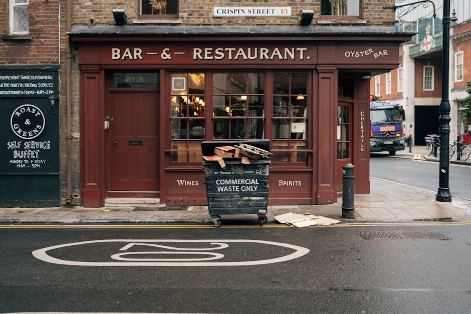Photograph of the exterior of a red-brick building housing a bar and restaurant, located on Crispin Street in Notting Hill. The shopfront features large glass windows with warm interior lighting visible inside. In front of the establishment, there is a black commercial waste bin labeled 'COMMERCIAL WASTE ONLY,' which is partially filled with flattened cardboard boxes and packing materials. The bin is situated on the sidewalk, slightly overlapping the curb. Nearby, a metal bollard is positioned on the pavement, while a black pole with decorative street lighting is located on the right side of the scene. To the left, a black signboard advertises a self-service buffet, and a black van is parked further down the street, suggesting typical urban loading and unloading activity. The scene captures a moment of waste disposal during a home relocation or furniture transport process, illustrating elements relevant to house removals and moving services in Notting Hill.