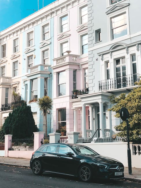 A black hatchback car parked on a narrow urban street in front of a row of white and pastel-coloured Victorian-style terraced houses with ornate facades, tall windows, and small balconies. The pavement features decorative iron railings and lush greenery, including a small palm tree and bushes. The scene depicts a typical setting for home relocation or furniture transport in Notting Hill, with the houses' architecture indicating a residential area suitable for house removals. The image illustrates the logistical considerations of moving services such as loading and unloading in tight, historic streets, highlighting the need for professional removals specialists like Man and Van Notting Hill to manage packing, moving, and transportation effectively.