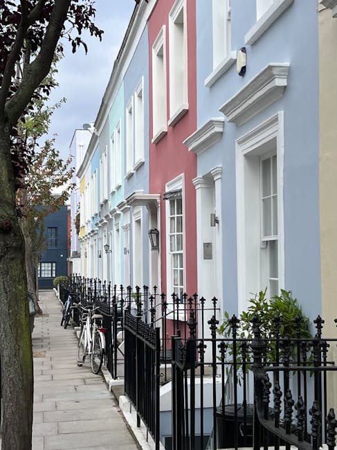 The image shows a row of colorful terraced houses on a narrow street in Notting Hill, London, with pastel-colored facades in shades of blue, pink, and yellow. The buildings feature white window frames and decorative architectural details, with small front gardens enclosed by black wrought iron fences. On the sidewalk, there are several bicycles leaned against the fences, and a large tree with green foliage partially visible on the left side. The pavement is made of light grey stone slabs, and the sky above is cloudy but bright. This residential scene captures the characteristic charm of the Holland Park Estate area, illustrating a typical setting where professional removals services by companies like Man and Van Notting Hill might operate during home relocation and furniture transport projects.