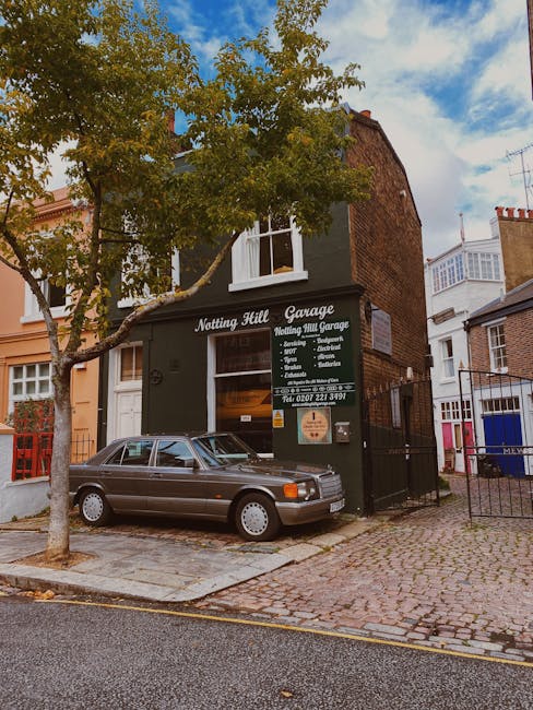 The exterior of Notting Hill Garage, a two-story brick building with a dark green facade, situated on a cobblestone street in Ladbroke Grove, Notting Hill. A vintage silver car is parked directly in front of the garage entrance, which displays signage for home relocation and furniture transport services. There is a large window on the first floor with white curtains, and a smaller window on the upper floor. A tall tree with green foliage partially obscures the building’s facade, casting shadows on the street below. The street is paved with cobblestones, and the sidewalk is made of concrete slabs. Adjacent buildings include residential terraced houses with red brick and pastel-colored exteriors, with some featuring decorative window trim. The sky is partly cloudy with patches of blue visible. This scene highlights the location’s proximity to residential areas often associated with house removals and moving services in Notting Hill, illustrating the typical urban environment for relocations serviced by companies like Man and Van Notting Hill.