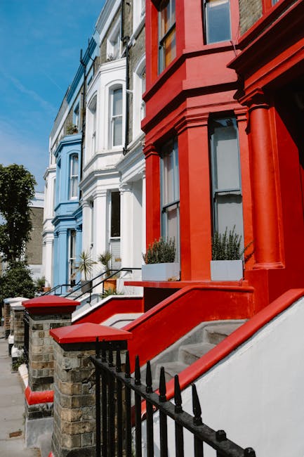 The image depicts a row of traditional terraced houses on Ladbroke Grove, Notting Hill, with a clear blue sky overhead. The buildings vary in colour, with one house painted pink featuring a decorative bay window and ornate architectural details, while adjacent properties are painted white with large sash windows and some with small decorative balconies. The façades include brickwork and painted render, with the pink house standing out prominently. The photograph is taken from the pavement, emphasizing the height and architectural features of the residences. The scene captures the typical Victorian-style terraced houses characteristic of Notting Hill, showing clean exteriors and well-maintained facades. This setting is relevant for visualising house removals, home relocation, and furniture transport, highlighting the importance of professional packing, loading, and moving logistics often supported by companies like Man and Van Notting Hill. Though no moving activity is visible, the image reflects the external environment where moving services facilitate interior packing, furniture transport, and building access for efficient home removals.