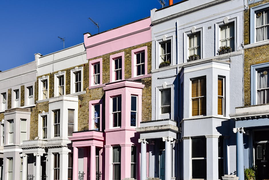 A row of colourful Victorian terraced houses in Notting Hill, London, painted in shades of blue, pink, yellow, and green. The buildings feature white bay windows and decorative mouldings, with some wi