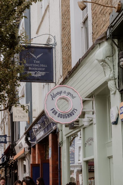 Photograph of a narrow street in Notting Hill with several shop signs visible, including one for 'The Notting Hill Bookshop' hanging from a metal bracket and another circular sign with a pink doughnut outline that reads 'Always a good FOR DOUGHNUTS'. The shopfronts are close together, featuring colorful facades with large windows and awnings. The scene is outdoors during daytime, with soft, natural lighting illuminating the buildings and signs. The photograph captures a busy, vibrant area known for its shops and cafés, with some pedestrians partially visible at the bottom of the frame. This urban setting reflects the charm of Notting Hill, which may be relevant in the context of house removals and moving services by Man and Van Notting Hill, who handle home relocations within the area.