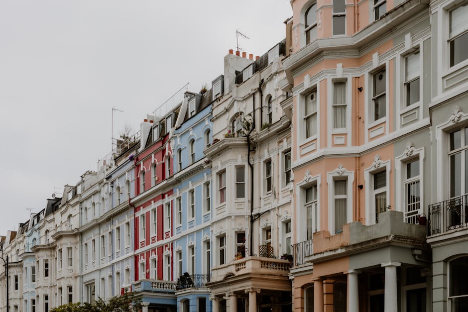 An exterior view of a row of Victorian-style terraced houses in Notting Hill, London, with pastel-colored facades in shades of pink, blue, and white, featuring large sash windows and decorative moldings. The buildings are multi-storey, with some featuring small balconies with black wrought iron railings, and are closely spaced along a narrow street. The sky is overcast, casting diffused natural light over the scene. In the foreground, the pavement runs parallel to the houses. This setting captures the typical residential architecture of Notting Hill, often associated with characterful home relocation and furniture transport projects, as handled by companies like Man and Van Notting Hill, especially for same-day emergency removals involving careful packing and loading processes for house moving services.