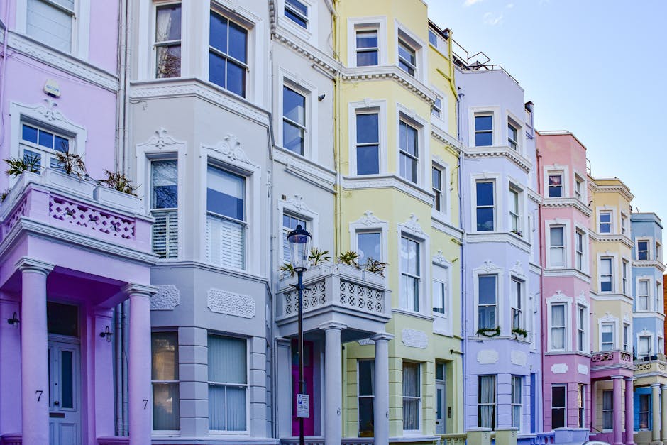 Photograph of a narrow street in Notting Hill with several shop signs visible, including one for 'The Notting Hill Bookshop' hanging from a metal bracket and another circular sign with a pink doughnut outline that reads 'Always a good FOR DOUGHNUTS'. The shopfronts are close together, featuring colorful facades with large windows and awnings. The scene is outdoors during daytime, with soft, natural lighting illuminating the buildings and signs. The photograph captures a busy, vibrant area known for its shops and cafés, with some pedestrians partially visible at the bottom of the frame. This urban setting reflects the charm of Notting Hill, which may be relevant in the context of house removals and moving services by Man and Van Notting Hill, who handle home relocations within the area.