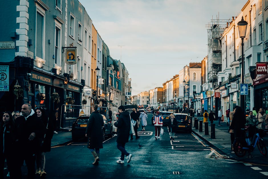 A busy street scene in Notting Hill during daylight hours, showing a mix of pedestrians walking along the pavement and crossing the road, with some individuals carrying shopping bags or engaged in conversation. Several parked and moving cars line the street, which features typical London-style terraced houses with shopfronts at ground level, including a visible pub named 'Duke of Wellington' on the left. The street is illuminated by classic lampposts, and some buildings are covered in scaffolding, indicating ongoing maintenance or renovations. The sky is partly cloudy, and the overall scene captures the lively atmosphere of this popular area, with moving vehicles and foot traffic, aligning with the themes of urban relocation and transportation support provided by Man and Van Notting Hill during home relocations or furniture transport processes.