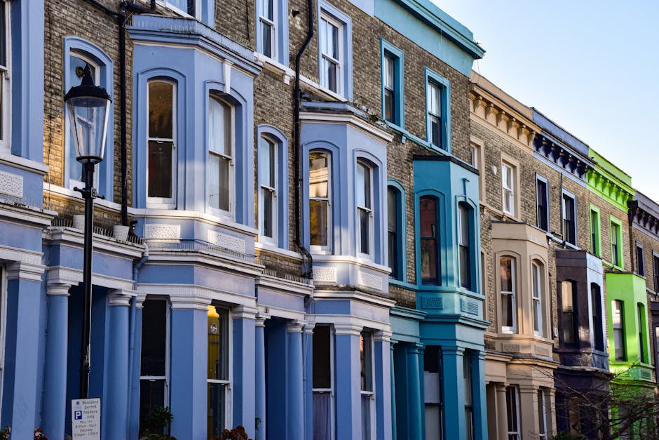 A row of Victorian terraced houses in Notting Hill with vibrant, pastel-colored facades, including shades of blue, beige, and bright green, featuring large sash windows and decorative architectural details. The buildings are three stories tall, with some windows open and reflecting the clear blue sky. In the foreground, there is a black street lamp and a metal parking sign on the pavement. Outside the houses, cardboard boxes, wrapped furniture, and packing materials are visible, indicating a home relocation process. Part of a delivery van is parked at the curb, with an open rear door loaded with additional boxes and moving blankets, suggesting furniture transport and packing for house removals. The scene captures a typical residential street, highlighting elements associated with moving and clearance services, as provided by Man and Van Notting Hill, in preparation for a move following local skip and disposal laws to avoid council fines.