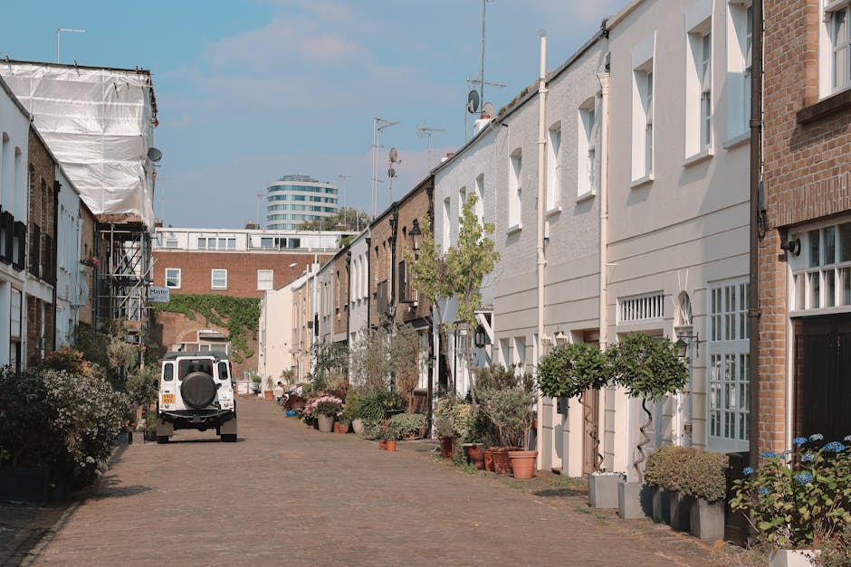A busy street scene in Notting Hill during daylight hours, showing a mix of pedestrians walking along the pavement and crossing the road, with some individuals carrying shopping bags or engaged in conversation. Several parked and moving cars line the street, which features typical London-style terraced houses with shopfronts at ground level, including a visible pub named 'Duke of Wellington' on the left. The street is illuminated by classic lampposts, and some buildings are covered in scaffolding, indicating ongoing maintenance or renovations. The sky is partly cloudy, and the overall scene captures the lively atmosphere of this popular area, with moving vehicles and foot traffic, aligning with the themes of urban relocation and transportation support provided by Man and Van Notting Hill during home relocations or furniture transport processes.
