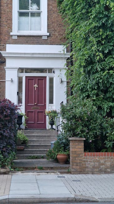 The image depicts the front entrance of a residential property in Notting Hill with a narrow staircase leading up to a maroon-colored wooden front door, which features a brass door knocker and mail slot. The entrance area is framed by white decorative trim and a white pediment above the door, set against a brick exterior wall. To the left side of the staircase, there are potted plants and small shrubs, with some pots placed on the steps and on the ground. On the right side, tall green foliage from a large bush or tree partially obscures the view, extending upward beyond the doorway. The scene is well-lit with natural daylight, highlighting the texture of the brickwork and the greenery. This setting illustrates common challenges encountered during home relocation or furniture transport in tight residential entrances, where careful planning and sometimes specialized moving equipment are required. Man and Van Notting Hill offers removals and moving services suited to such properties, especially those with narrow doorways and stairs.