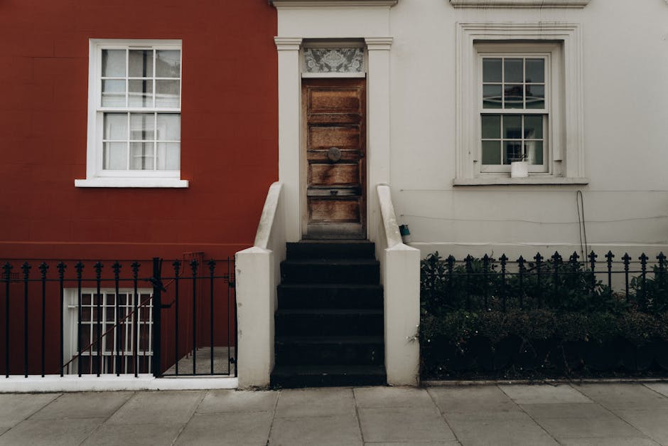A photograph of a residential building in Notting Hill showing a narrow doorway between two walls, one painted in a dark red hue and the other in white. The doorway is accessible via a small set of black-painted concrete stairs with white concrete sidewalls. There are white-framed sash windows above and beside the entrance, with one window on each side of the doorway, both having multiple panes and white trim. The red wall on the left has a white window with multiple panes, positioned below ground level, and a black metal fence runs along the sidewalk in front of this lower window and the steps, with a gate visible on the left side. The white wall on the right features a window with a small white ledge and a terracotta pot placed on it. The building exterior is illuminated by natural daylight, and there are no vehicles or people visible in the image, capturing a moment relevant to home relocation or furniture transport in the context of staircase and narrow-doorway moving challenges in Notting Hill, as handled by Man and Van Notting Hill.