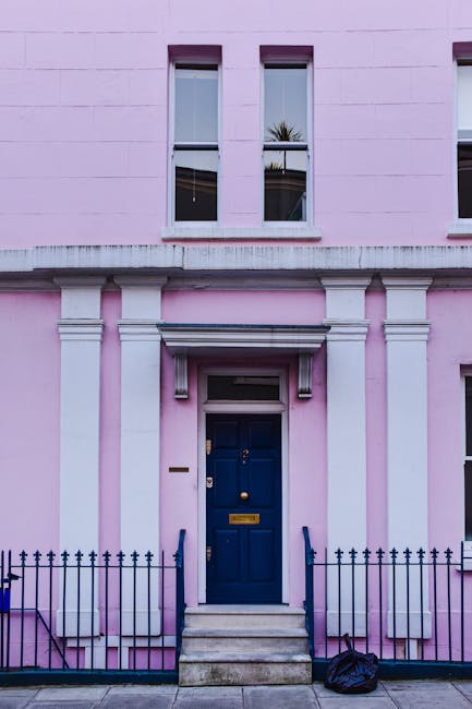 The image depicts the front entrance of a residential property in Notting Hill with a narrow staircase leading up to a maroon-colored wooden front door, which features a brass door knocker and mail slot. The entrance area is framed by white decorative trim and a white pediment above the door, set against a brick exterior wall. To the left side of the staircase, there are potted plants and small shrubs, with some pots placed on the steps and on the ground. On the right side, tall green foliage from a large bush or tree partially obscures the view, extending upward beyond the doorway. The scene is well-lit with natural daylight, highlighting the texture of the brickwork and the greenery. This setting illustrates common challenges encountered during home relocation or furniture transport in tight residential entrances, where careful planning and sometimes specialized moving equipment are required. Man and Van Notting Hill offers removals and moving services suited to such properties, especially those with narrow doorways and stairs.
