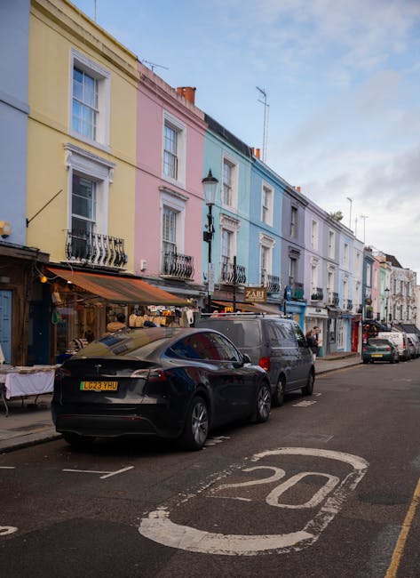 A row of colorful terraced houses on a street in Notting Hill with pastel facades in shades of yellow, pink, blue, and white, each with decorative window frames and small balconies. Parked cars, including a black sports car and a grey van, are positioned along the curb in front of the residences. The street features a marked speed limit of 20 mph painted on the asphalt and a narrow sidewalk with display tables extending from shopfronts under striped awnings. A street lamp is visible between two of the buildings, and the sky is partly cloudy with patches of blue. This scene illustrates a typical residential area suitable for home relocation or furniture transport services, with vehicles positioned properly for loading or unloading. Man and Van Notting Hill's removal service may be involved in the logistics of moving items through this street, supporting packing and moving processes.